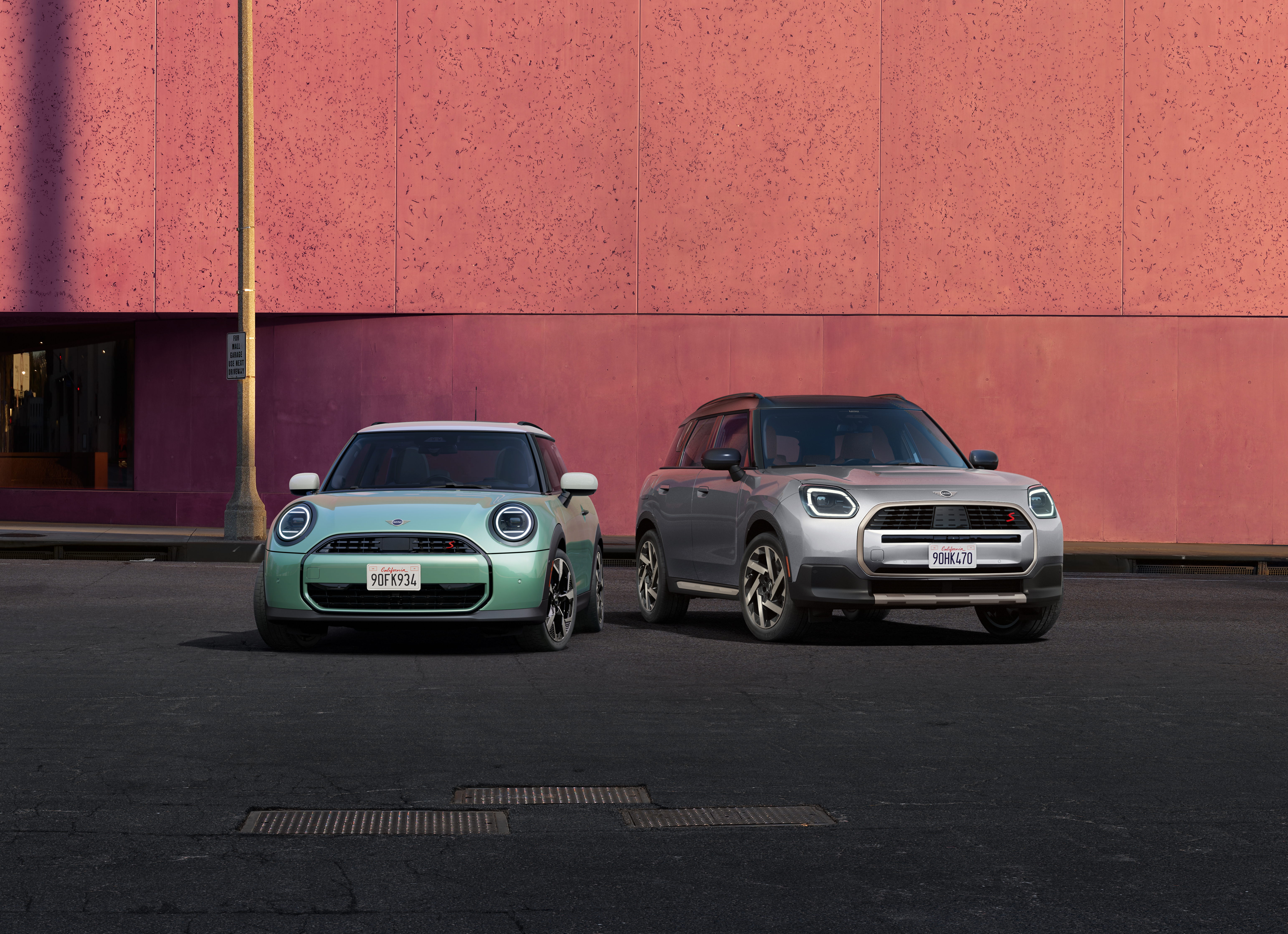 Front-view of the 2025 MINI Cooper S 2 Door in Ocean Wave Green and 2025 MINI Countryman S ALL4 in Melting Silver III parked side by side in front of a salmon-colored concrete building.