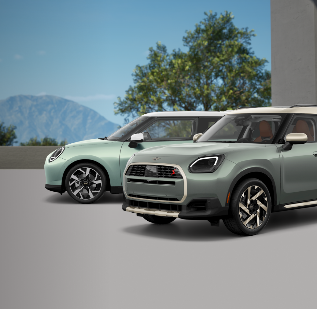 Angled view of a Countryman S ALL4 in Smokey Green Metallic parked on a rooftop with a MINI Cooper S 2 Door parked behind it with a mountain and trees in the background.
